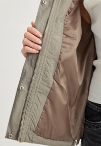 Ljusgrå jacka med en strukturerad utsida och slät, beige satinfoder. Dragkedjestängning med metallbeslag och tryckknappsdetaile.