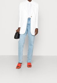 Vit kavaj över en ljus skjorta med knappar, ihop med ljusblå jeans och röda platta sandaler. Svart handväska i handen, med minimalistisk design.