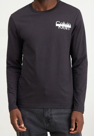 Mężczyzna ubrany w czarną koszulę Calvin Klein z długim rękawem i białym logo na piersi, w parze z ciemnoszarymi dżinsami.