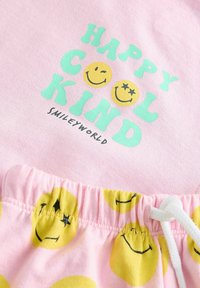 Rosa tyg med texten "HÄRLIG GERSAJ" i grön färg. Den elastiska midjebandet har ett mönster av gula smiley-ansikten med en vit dragsko.