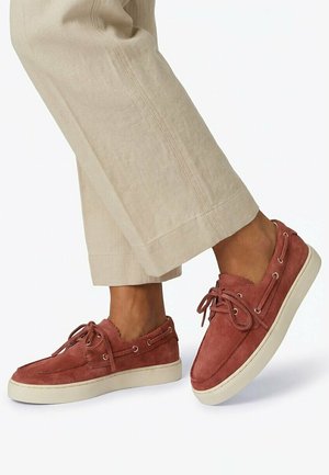 Pieds chaussés de baskets en daim couleur rouille à lacets avec semelles blanches, associées à un pantalon beige à jambes larges sur fond blanc.