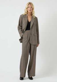 Grijze op maat gemaakte blazer met notchrevers en enkele knopen sluiting, gecombineerd met een broek met wijde pijpen. Ggedragen over een zwarte top, van gestructureerde stof.