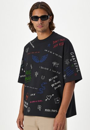 Homme portant un t-shirt noir oversize avec un texte manuscrit coloré et des griffonnages, pantalon beige et lunettes de soleil noires, regardant légèrement vers la gauche.