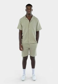 Hellgrünes Kurzarm-Hemd mit Knopfleiste und passenden Shorts, aus strukturiertem Stoff. Getragen mit weißen Socken und Schuhen.