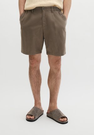 Man draagt bruine casual korte broek en grijze open teen slippers, staat met handen in zijn zakken tegen een effen achtergrond.