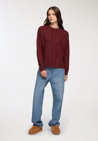 Maglione borgogna a lavorazione a treccia con scollatura rotonda, abbinato a jeans denim blu larghi e pantofole in camoscio marroni. Design semplice e testurizzato.