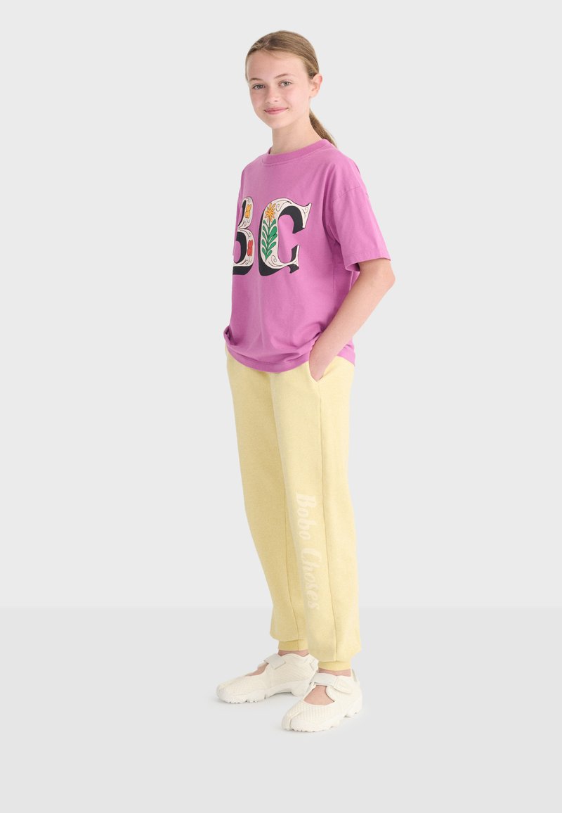 Fille debout, les mains dans les poches, portant un t-shirt graphique violet, un pantalon de survêtement jaune avec texte sur le côté, et des chaussures de sport blanches sur un fond clair.