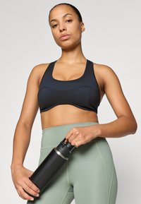 Top deportivo negro con escote redondeado y tirantes anchos, combinado con leggings de talle alto en verde claro. Sosteniendo una botella de agua negra.