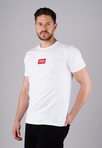 Mann mit kurzen Haaren und Bart, der ein weißes T-Shirt mit rotem "PICALDI"-Logo trägt. Er steht mit einer Hand in der Tasche seiner schwarzen Hose vor einem grauen Hintergrund.