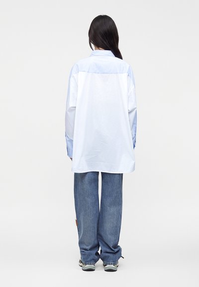 Femme aux longs cheveux foncés, portant une chemise blanche oversize avec des manches rayées bleues et un jean bleu ample, debout de dos.