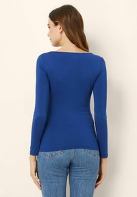 Top albastru cu mânecă lungă și decolteu în formă de semicerc, realizat dintr-un material elastic, asortat cu blugi din denim de un albastru deschis, văzut din spate.
