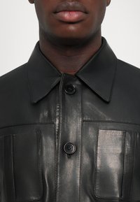 Tuvumā redzams cilvēks, kurš valkā melnu ādas jaku ar izteiktu apkakli un divām pogām aiztaisāmām krūšu kabatām.