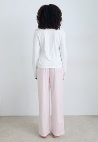 En person med lockigt hår står med ryggen mot kameran, iklädd en vit långärmad skjorta och rosa vida byxor mot en ljus bakgrund.