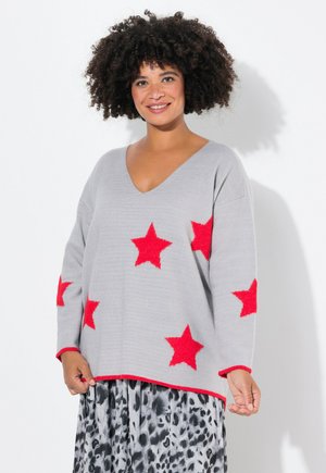 Maglione grigio con scollo a V e maniche lunghe, caratterizzato da stelle rosse e un bordo rosso lungo l'orlo. Indossato sopra una gonna a fantasia.