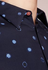 Mörkblå skjorta med ljusblå cirkulära mönster och en enda röd knapp nära kragen, som bärs nära en ljus hudton på nacken.