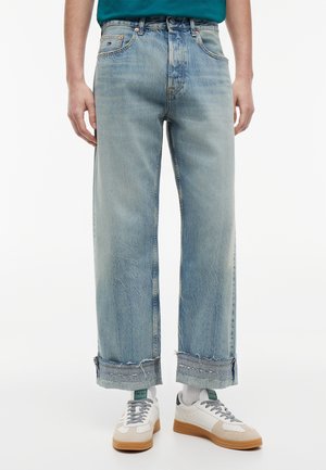 Osoba nosząca podwinięte niebieskie jasne dżinsy, białe skarpetki i białe trampki z gumowymi podeszwami, stojąca na jednolitym tle.