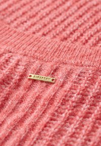 Sweter dzianinowy w miękkim koralowym kolorze z teksturowanym wzorem. Metalowy znacznik z napisem "SUPERDRY" w kolorze złotym.