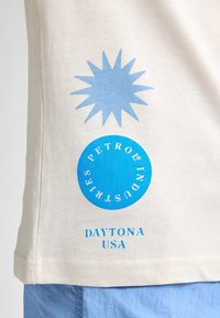 Camiseta de algodón beige con un graphic de estallido de sol azul claro y un logotipo circular con "PETROL INDUSTRIES" y "DAYTONA USA" en texto azul.