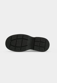 Semelle de chaussure en caoutchouc noir avec des motifs de bande de roulement rectangulaires et courbés sur un fond gris clair.