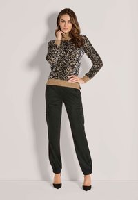 Frau, die in einem turtleneck Pullover mit Leopardenmuster, schwarzen Cargo-Hosen mit elastischen Bündchen und schwarzen spitzen High Heels vor einem einfarbigen Hintergrund posiert.