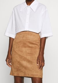 Chemise blanche à manches courtes et à boutons avec un large col, associée à une jupe crayon en daim beige. La jupe présente une texture lisse et des coutures discrètes.