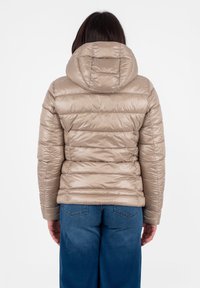 Giacca imbottita beige con sezioni trapuntate e cappuccio. Texture liscia e lucida, con polsini elastici. Indossata con jeans blu, vista da dietro.