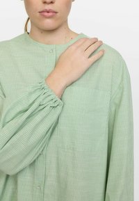 Cămășă butonată cu dungi verde deschis, dintr-un material ușor, cu un guler rotund, cu manșete răsfrânte și o singură buzunar pe piept.