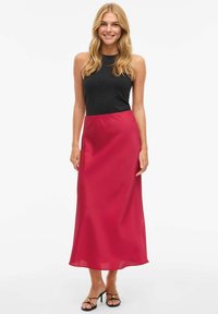 Svart ermeløs topp kombinert med et flytende, midjelangt, lyse rødt satengskjørt. Modellen har på seg svarte, remmede hæler med gullaksenter.