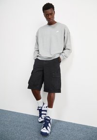 Sweatshirt cinza com um logotipo branco, calções de carga pretos e sapatilhas azul-marinho com detalhes brancos. Meia visível acima do tornozelo. Design simples.