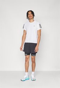 Weißes Sport-T-Shirt mit schwarzen Streifen an den Ärmeln, kombiniert mit schwarzen Shorts und weißen Sneakers mit blauen Akzenten. Einfaches und sportliches Design.