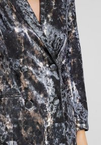 Blazer de corte cruzado en un patrón texturizado en negro y plata. Presenta un cuello de muesca, botones prominentes y un acabado suave y aterciopelado.