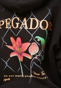 Black hoodie featuring a floral design in red and pink, with a colorful bird and stylized text on the front. The background has a chain-link pattern.