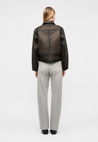 Γυναίκα με ίσια ξανθά μαλλιά, που φοράει μαύρο δερμάτινο μπουφάν και ανοιχτό γκρι τζιν ψηλόμεσο με ίσια μπατζάκια, στέκεται γυρισμένη με την πλάτη.