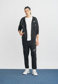 Fekete sportos melegítő, cipzáros kapucnival zöld díszítéssel és texturált rétegekkel, hozzáillő melegítőnadrággal párosítva. Fehér sportcipő.