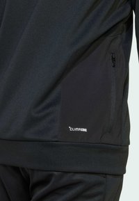 Top sportivo nero con una texture liscia, dotato di una tasca con zip sul lato sinistro e logo "CLIMACOOL" in bianco sull'orlo.