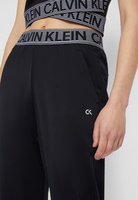 Czarne dresy Calvin Klein mają elastyczny pas z wzorem w paski, boczne kieszenie oraz małe białe logo CK na lewej udzie.