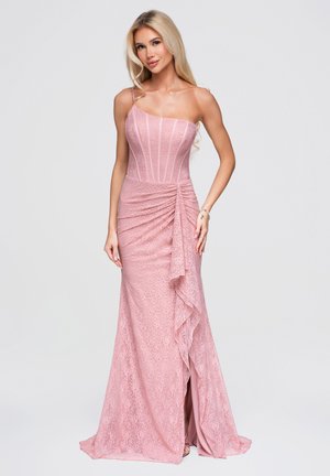 Femme aux longs cheveux blonds portant une robe en dentelle rose asymétrique avec des détails froncés et une fente sur le devant, debout devant un fond uni.