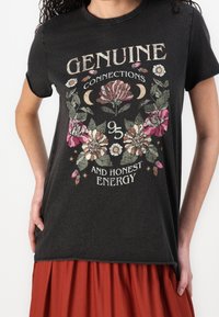 Zwarte katoenen t-shirt met korte mouwen, bloemige grafische print in roze en wit, met de tekst "Echte Verbindingen" en "Eerlijke Energie."