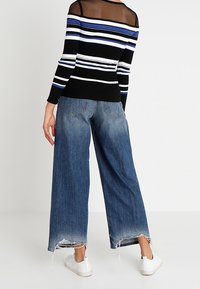 Gestreifter Pullover in Schwarz, Weiß und Blau; kombiniert mit weit geschnittenen blauen Jeans mit ausgefranstem Saum. Weiße Sneakers sichtbar.