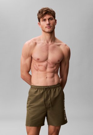 Homme musclé debout, les mains derrière le dos, portant un short de bain Calvin Klein vert olive, sur fond gris clair uni.