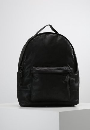 Schwarzer Leder-Rucksack mit vorderer Reißverschlusstasche und Tragegriff oben, auf einer weißen Oberfläche stehend vor einem schlichten hellen Hintergrund.