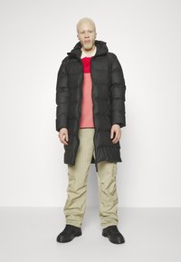 Zwarte pufferjas met capuchon, ritssluiting en bungee-detail. Gelaagd over een rood en crèmekleurig trui, gecombineerd met beige broek en zwarte laarzen.