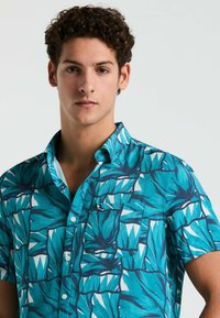 Korte mouwen shirt met een turquoise achtergrond en een gedurfde bladenpatroon in blauw en wit, knoopsluiting aan de voorkant en een borstzak.