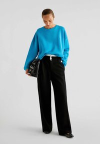 Turquoise oversized sweatshirt, zwarte wijde broek, zwarte tas met zilverkleurige accenten, en zwarte flats met gespikkelde details.
