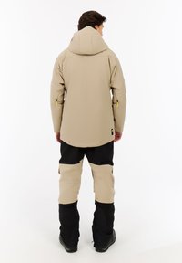 Beige wasserfeste Jacke mit Kapuze, kombiniert mit schwarzen Hosen mit einem beigen Streifen. Die Jacke hat eine glatte Textur und einen Reißverschluss an den Ärmeln.