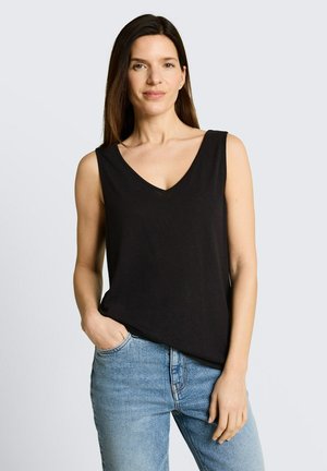 Frau mit langen braunen Haaren, die ein schwarzes ärmelloses V-Ausschnitt-Top und blaue Jeans trägt, steht mit einer Hand in der Tasche vor hellem Hintergrund.