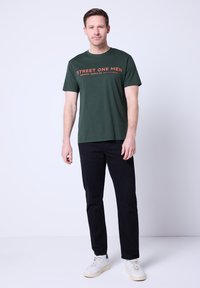 Zielona bawełniana koszulka z krótkim rękawem z pomarańczowym napisem: "STREET ONE MEN ORIGINAL SUPPLY CO." W połączeniu z czarnymi spodniami i białymi trampkami.