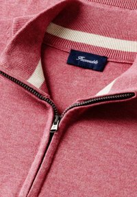 Gros plan sur le col d'un pull en tricot rose avec une bande blanche, une fermeture éclair métallique partiellement ouverte, et une étiquette de marque bleu foncé à l'intérieur.