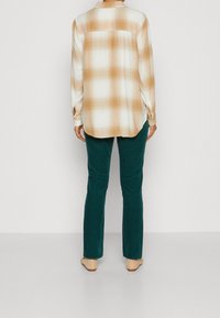 Camisa larga a cuadros en beige y blanco con mangas remangadas, combinada con pantalones azul oscuro y calzado claro. Tela suave y corte relajado.