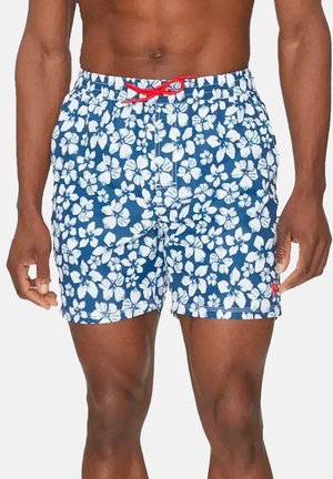 Short de bain bleu avec imprimé fleur d’hibiscus blanche et cordon rouge, porté par une personne debout à la peau foncée.